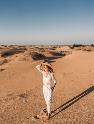 stylish beautiful woman walking in desert sand in white outfit wearing straw hat on sunset, travel safari on vacation